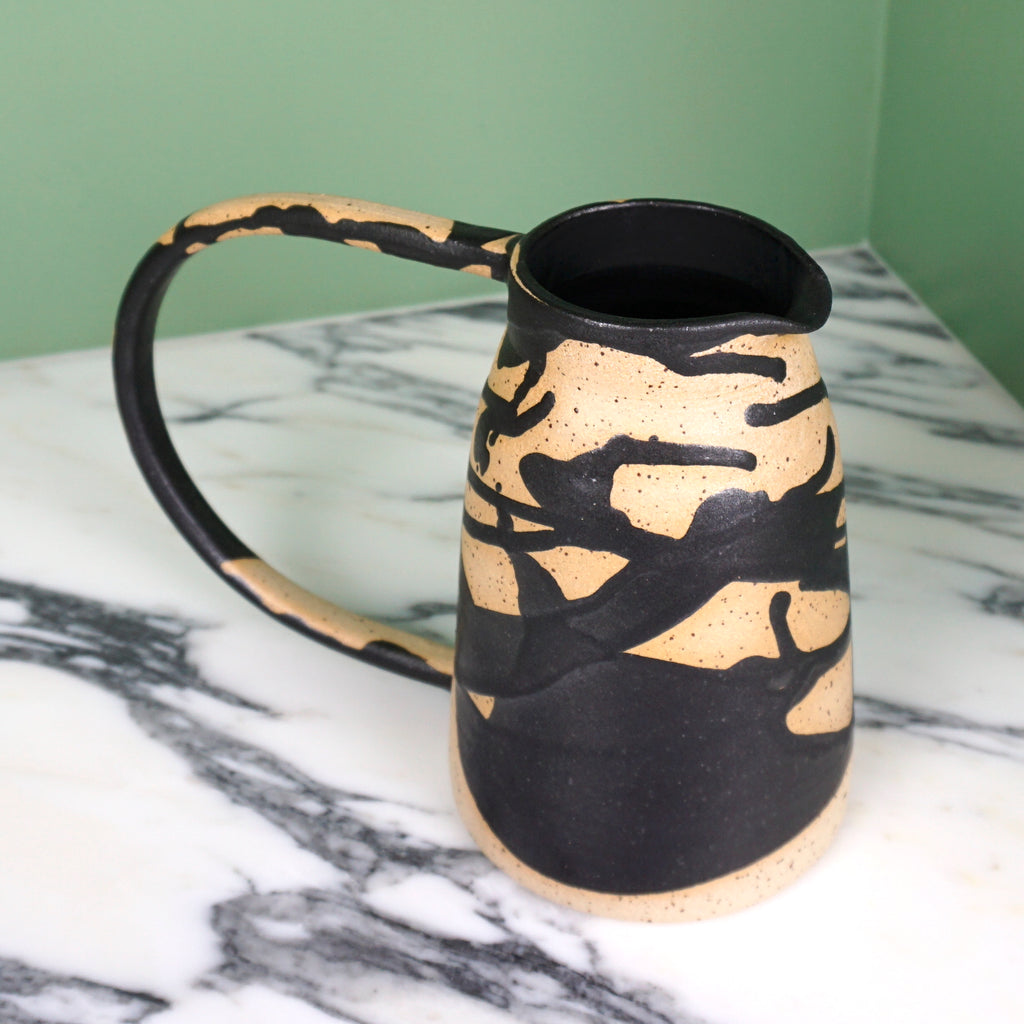 stoneware jug with a black drip glaze and an oversized handle. on a marble table with a green background. 