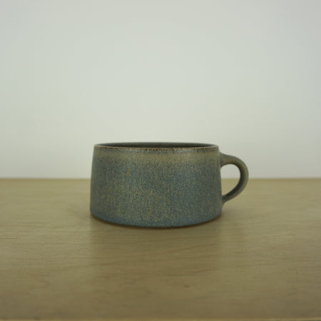 handmade ceramic mug by aku ceramics. simple shape in a blue green glaze. on a wooden table with a white wall. 