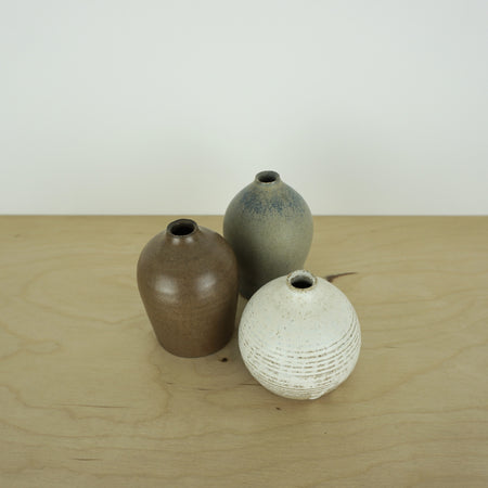 three mini handmade ceramic vases by Aku ceramics on a table. one white, one blue and one brown. 