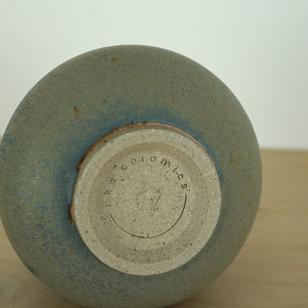 small handmade blue green cup on a wooden table and a white background. on its side so you can see the base and the makers mark of Aku Ceramics. 