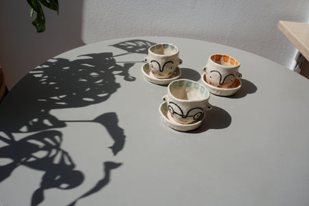 a group of three small pinch pot planters each with its own saucer, sitting on a table. one green, one orange and one blue. all with faces painted on. 