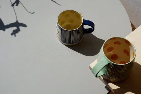 a green mug on a shelf and a blue mug on a table next to it. 