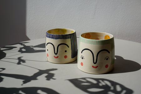 a green mug and a blue mug by beci callow both with faces hand painted on to them. 