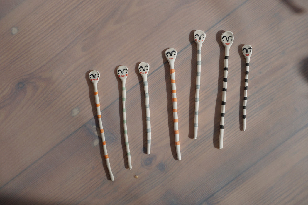 a group of multicoloured striped salt spoons lying on a piece of wood. all with faces painted on the end by Beci Callow