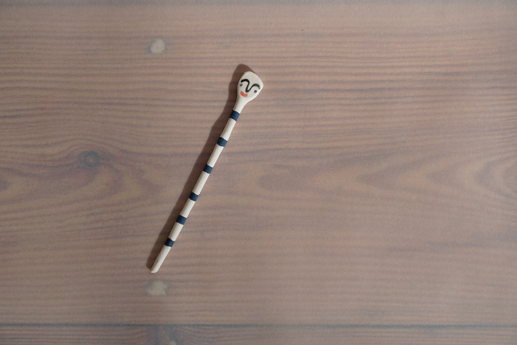 a ceramic salt spoon lying on a table in the sunshine. it has black stripes and a face painted on the end. 