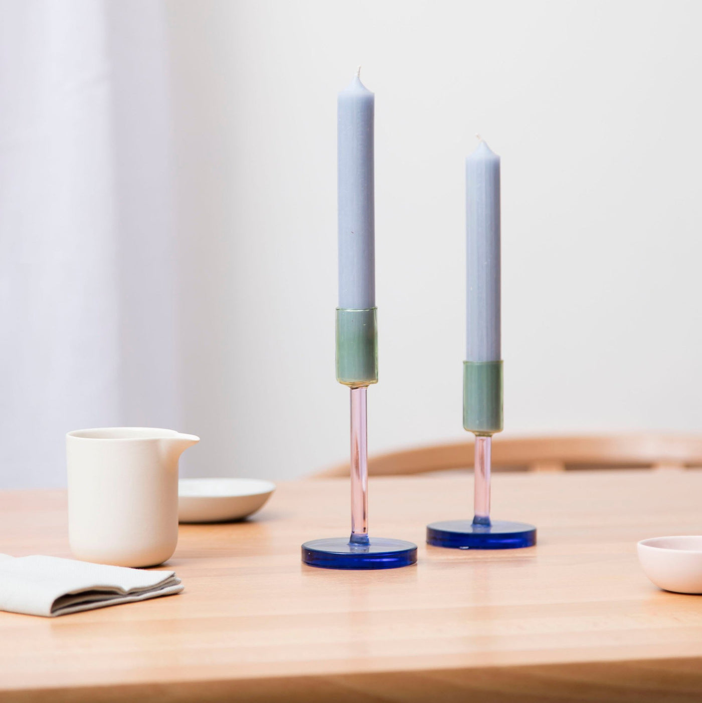 two glass candlestick holders on a table with a jug and salt dish 