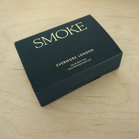 a box for a soap lying on the table by the brand Evermore London. the box is black and has the word Smoke written on it in gold lettering. 