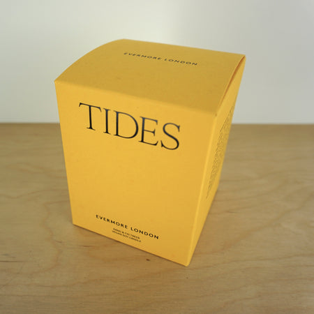 Evermore London candle box on a wooden table. the box is yellow with black lettering that says tides. 