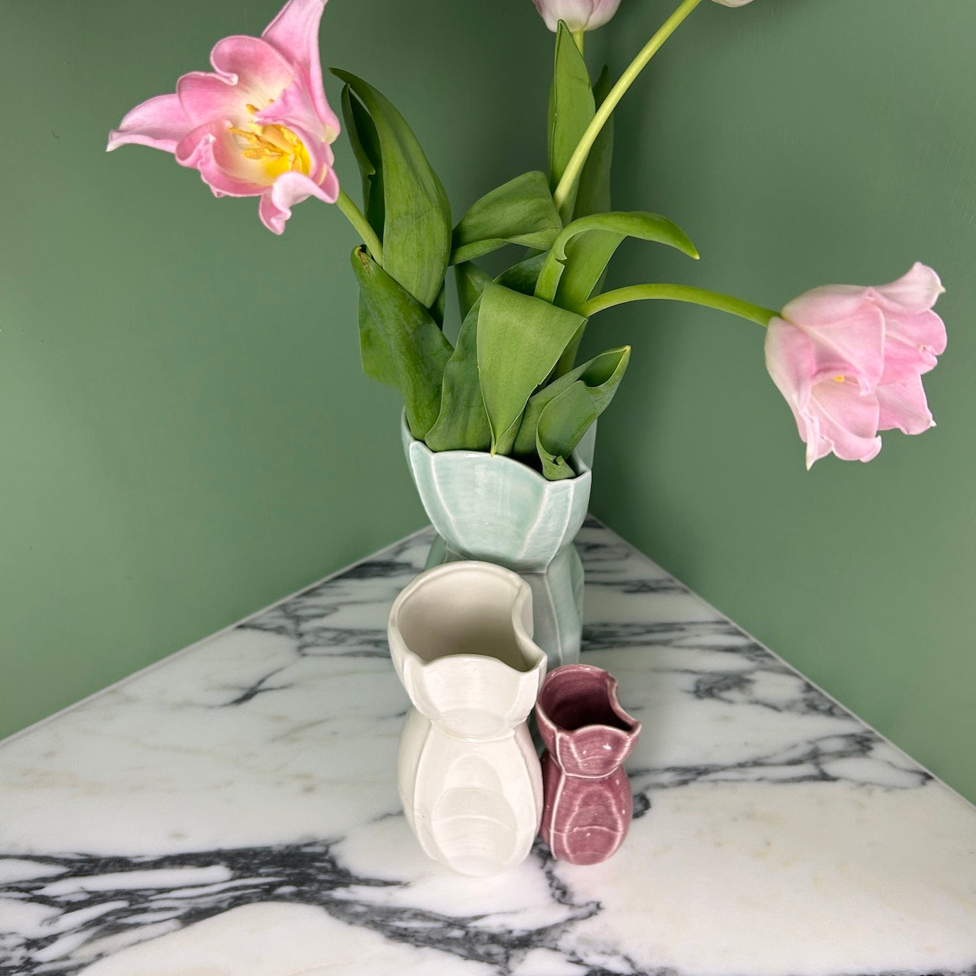 pink tulips in a green vase next to a white vase and a red vase on a marble table. 