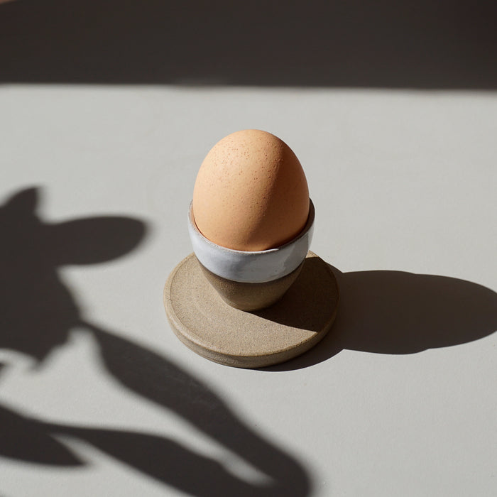 a stoneware egg cup, half glazed in white in the sunshine with an egg in it. 