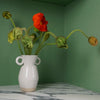 a white vase with two lugs at the top. sitting on a marble table with a bunch of red poppies. 