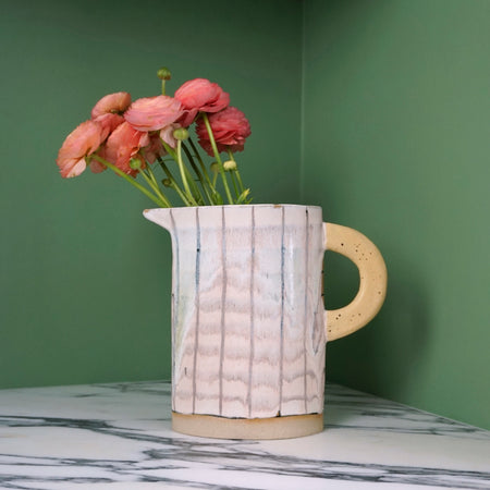 handmade jug with yellow handle and a bunch of pink flowers on a marble table with a green background. 