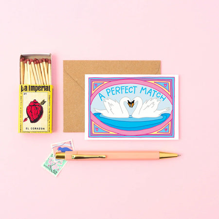 a card with two swans on a pond that reads A Perfect Match. next to a pen and a match box on a pink table. 