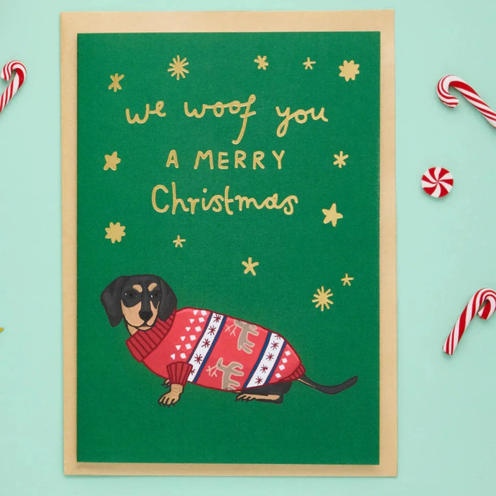 an illustrated christmas card of a sausage dog in a knitted jumper with gold letters reading We woof you a merry Christmas. 