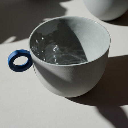 a round grey mug by anna jones ceramics sitting in the sunlight with a blue handle. 