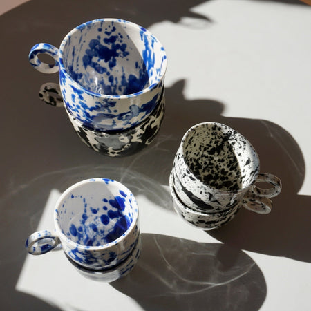 a group of two stacked round mugs and four stacked espresso cups on a table in the sunlight. all with a blue or black splatter glaze. 