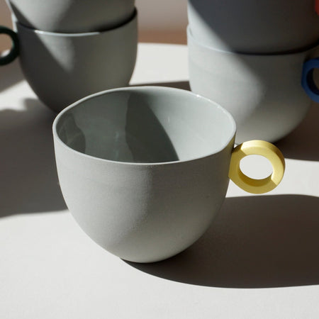 in the foreground a grey mug with a yellow handle. four mugs are stacked behind with different coloured handles. 