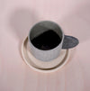 a grey espresso cup with a darker grey handle on a sand coloured saucer. sitting on a table. 