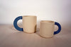 a large mug and a smaller mug. both sand coloured clay with a half moon blue denim handle. 