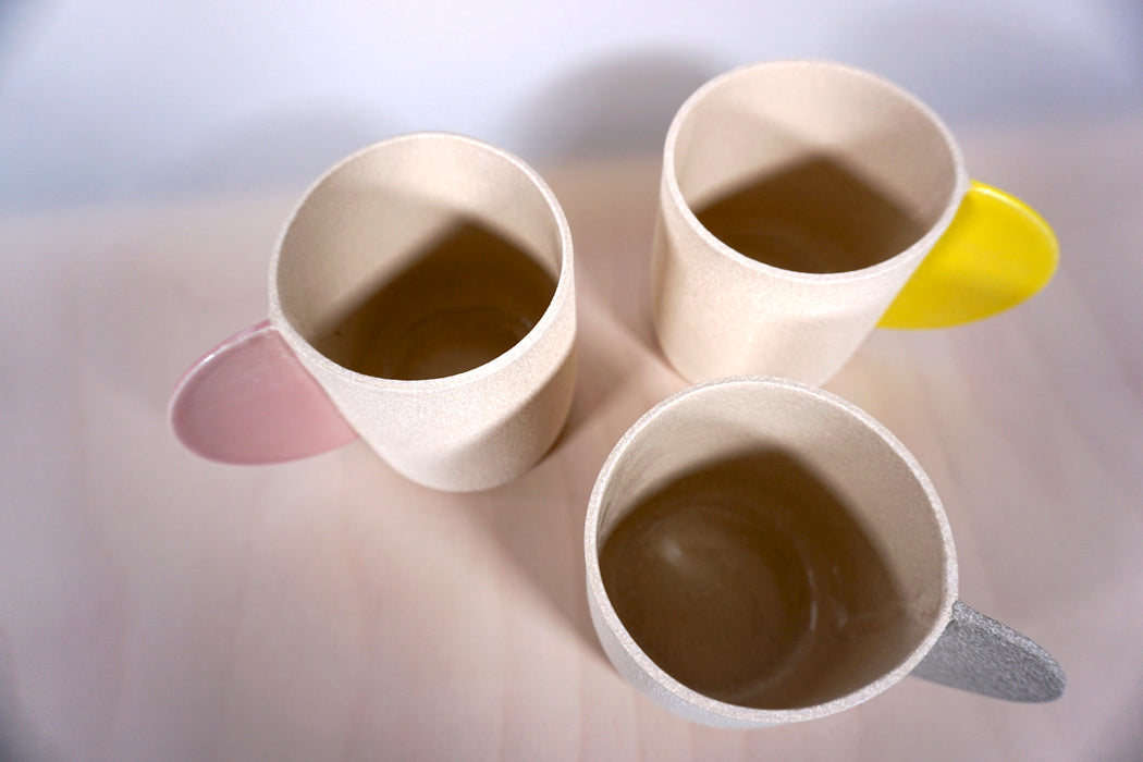 three sand coloured mugs in textured stoneware by Brutes Ceramics. one yellow handle, one pink handle and one grey handle. 