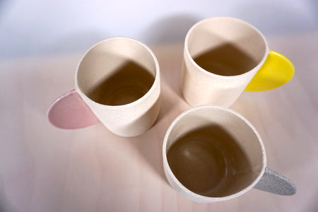 three sand coloured mugs in textured stoneware by Brutes Ceramics. one yellow handle, one pink handle and one grey handle. 