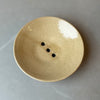 an oatmeal coloured soap dish with three drainage holes on a grey table. 