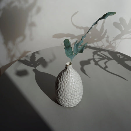 a white vase on a blue grey table with a single leaf stem in the vase. 