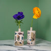 two collaged bud vases on a marble table with a green back ground. one has a yellow flower and one has a blue flower. 