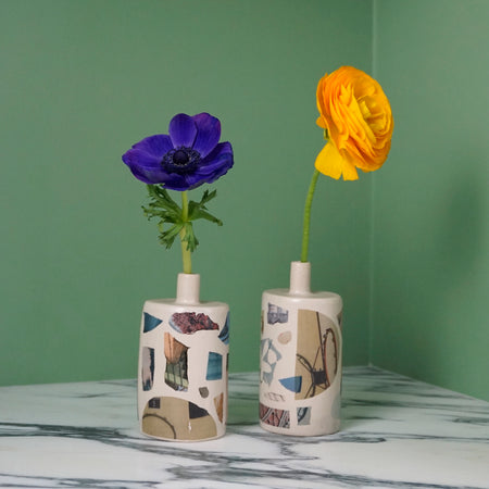 two collaged bud vases on a marble table with a green back ground. one has a yellow flower and one has a blue flower. 